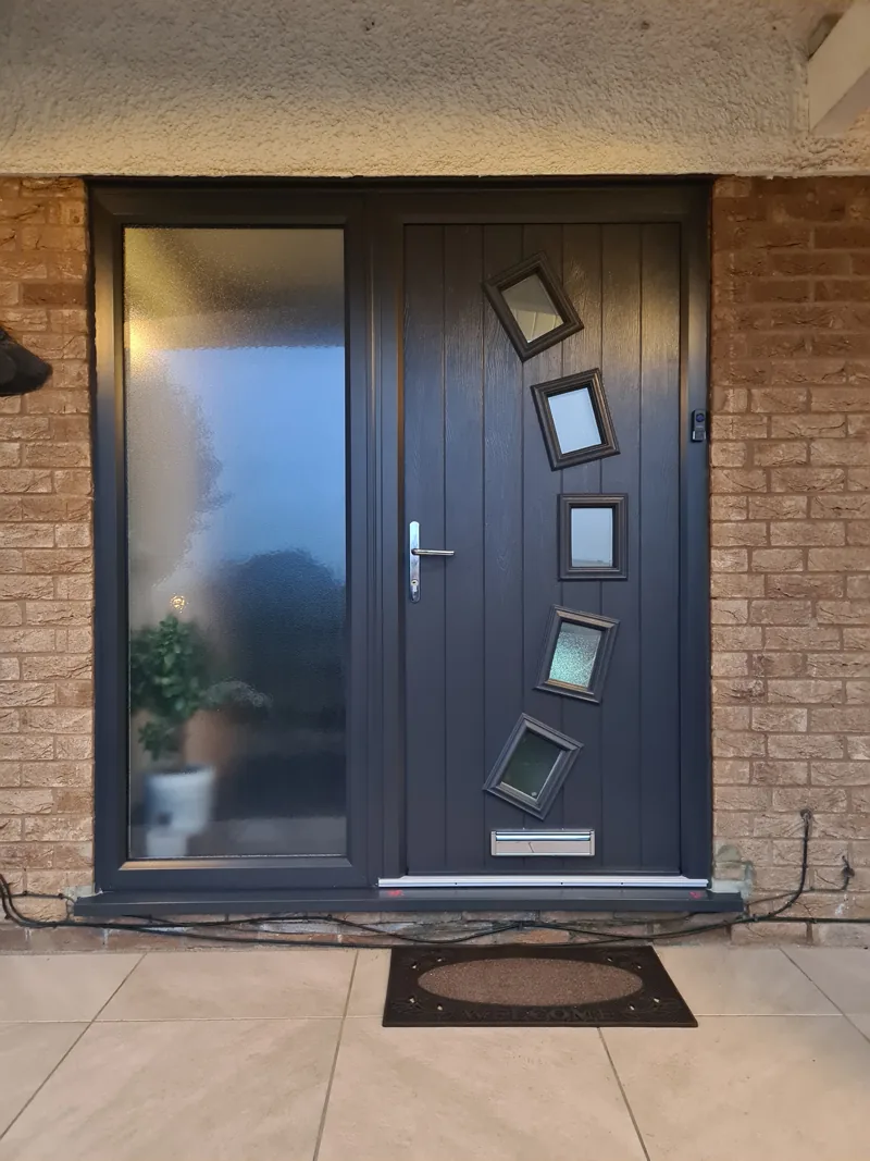 An anthracite grey composite front door with four diamond-set glazed panes, photographed at dusk with the porch light on and a mat at the threshold.
