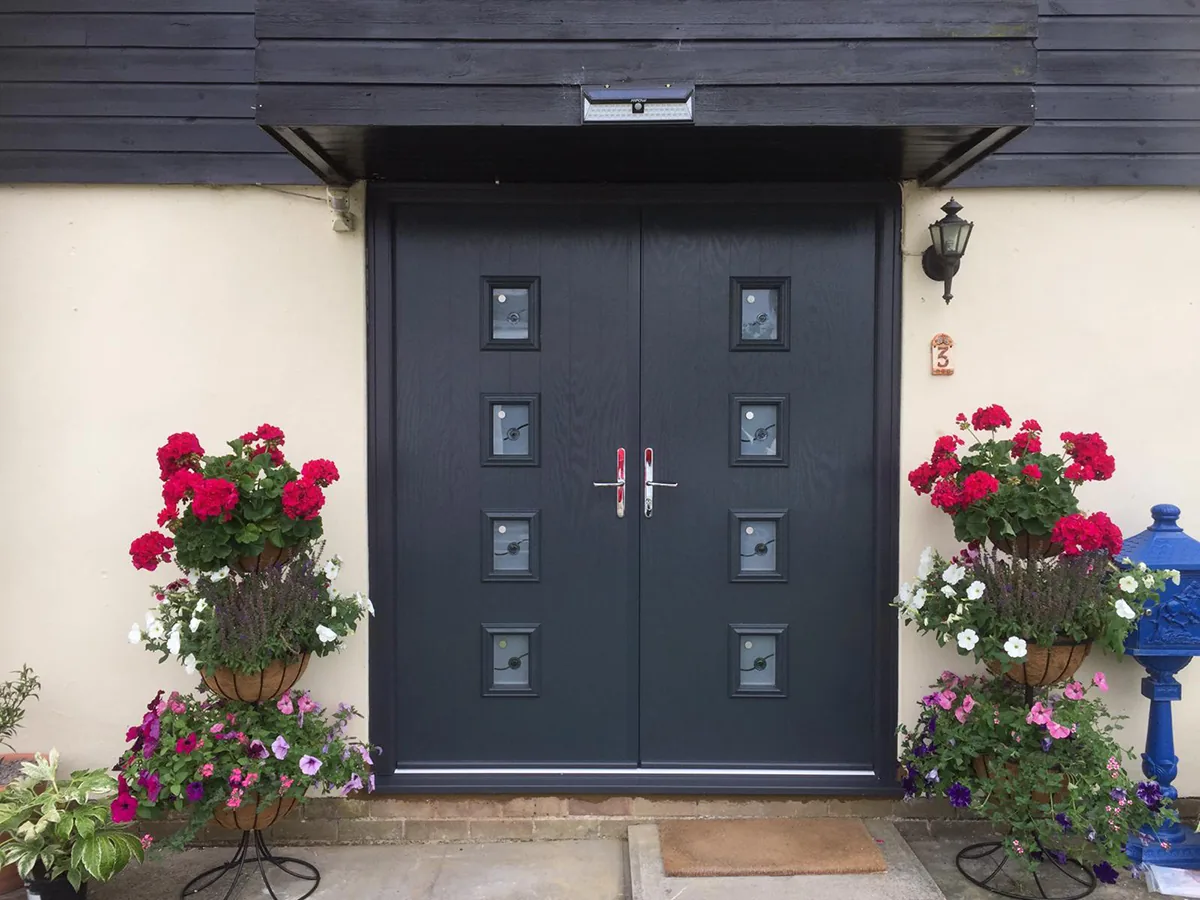 An anthracite grey double composite front door with six square glazed panels, fitted under a black timber canopy in a rendered porch, with geraniums in hanging baskets either side and a stone step at the threshold.