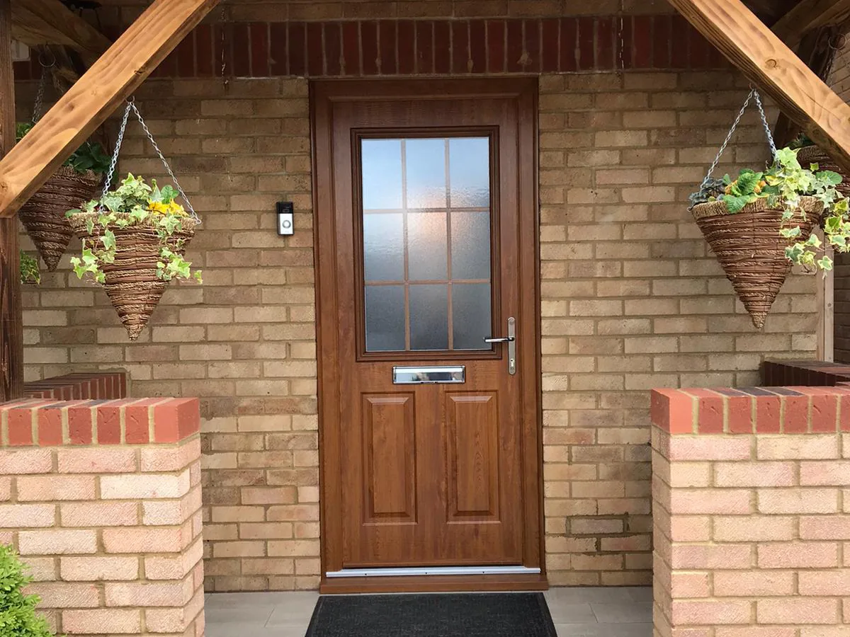 An oak-effect woodgrain composite front door with a glazed upper panel and a chrome letterplate, in a light brick porch between two brick piers with hanging baskets.