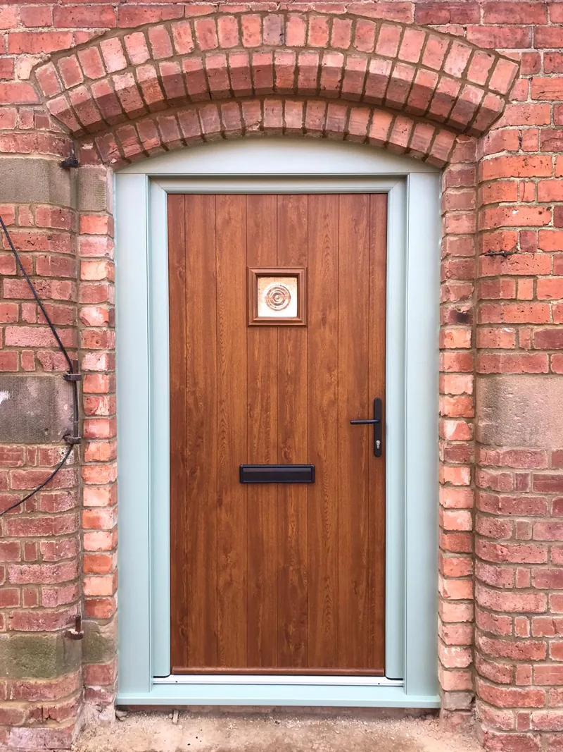 An oak-effect woodgrain composite front door with a brass knocker, fitted under the brick arch of a porch with a tiled step.