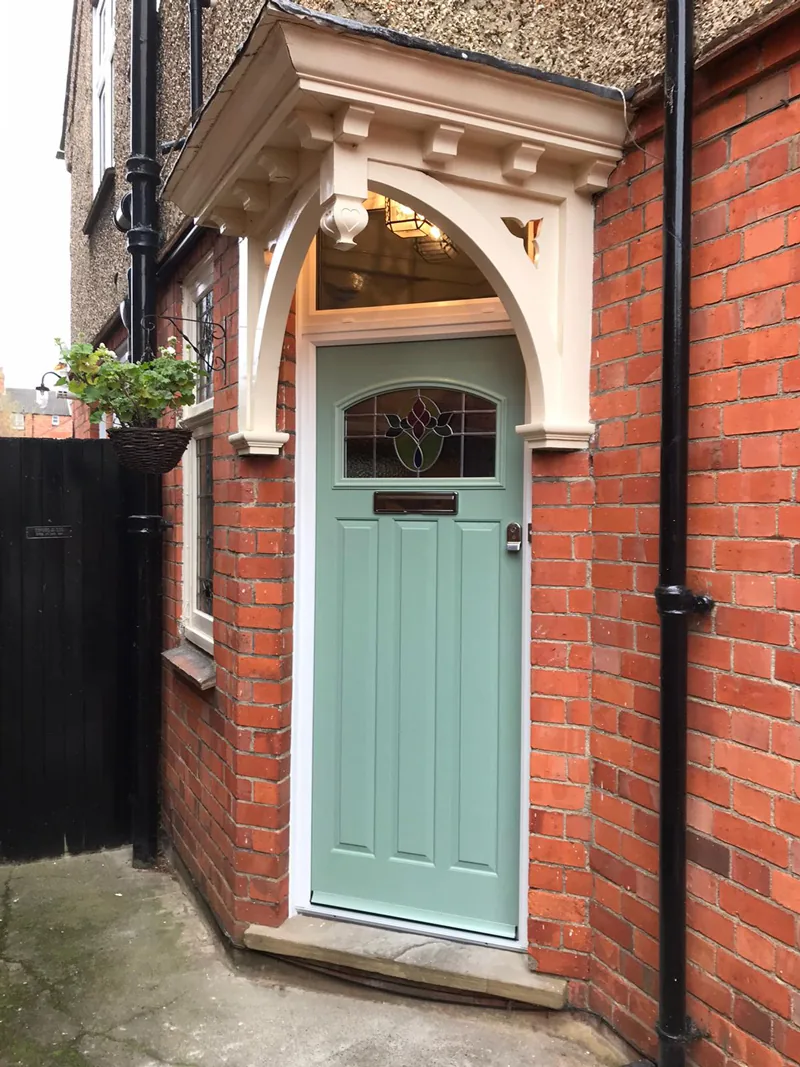 A sage green composite front door with a leaded stained-glass light and a brass letterplate, fitted into the arched opening of a Victorian red-brick porch above a stone step.