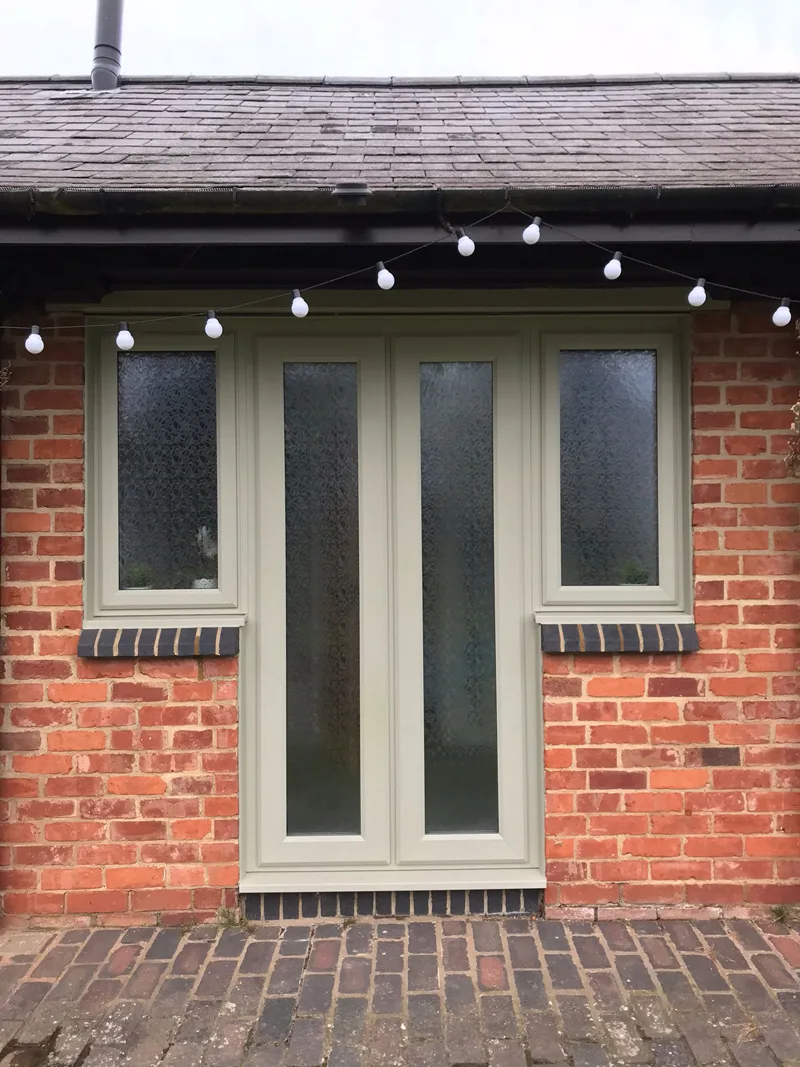 Sage green French doors with obscured glazing and matching side windows, in a red-brick elevation with festoon lights strung above them and a brick paved patio in front.