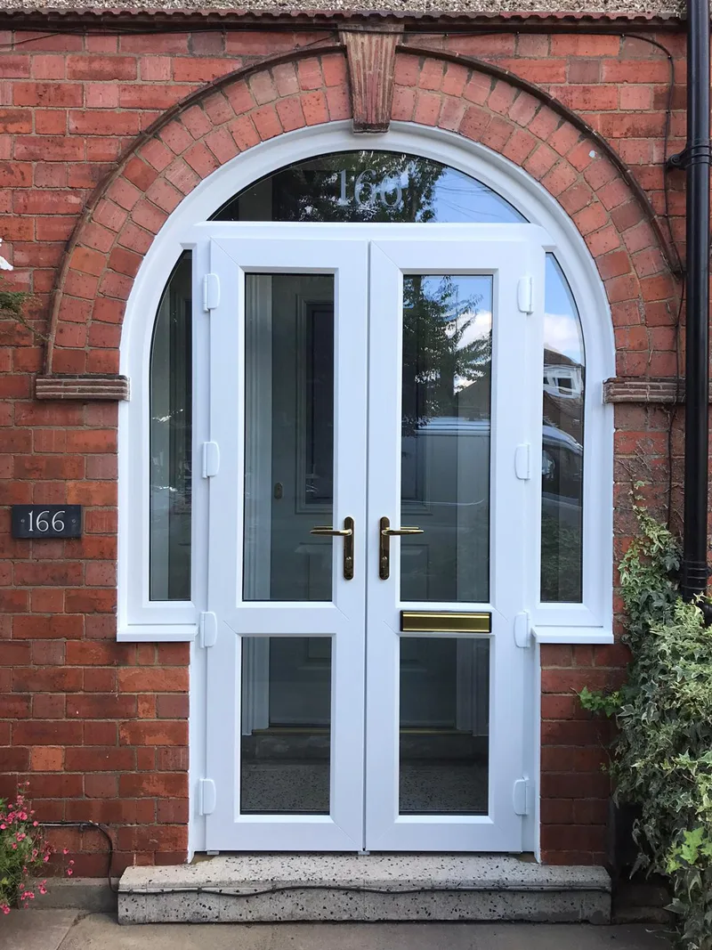 A pair of white uPVC doors with brass handles and a letterplate, set under an arched fanlight in a red-brick opening beside a house number plaque reading 166.
